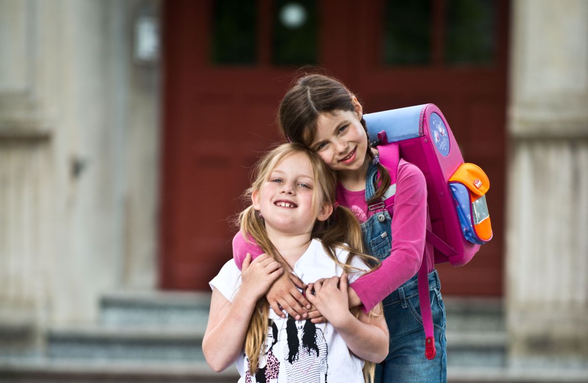Zwei kleine Schulmädchen mit Tornister auf dem Rücken umarmen sich freundschaftlich.
